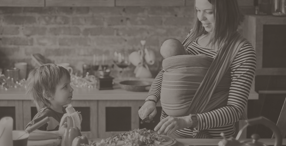 gray-scale image of a mother preparing a meal, she is wearing child carrier with a small baby in it and another child is watching her