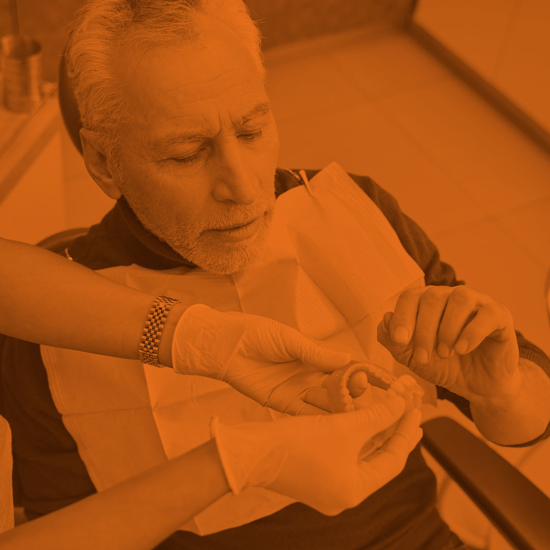 orange-scale image of a man sitting in a dental chair looking at a pair of dentures with a dentist in the foreground holding the dentures