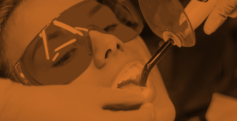 orange-scale image of a person wearing safety glasses with a dentist's hands holding a dental tool in the person's mouth