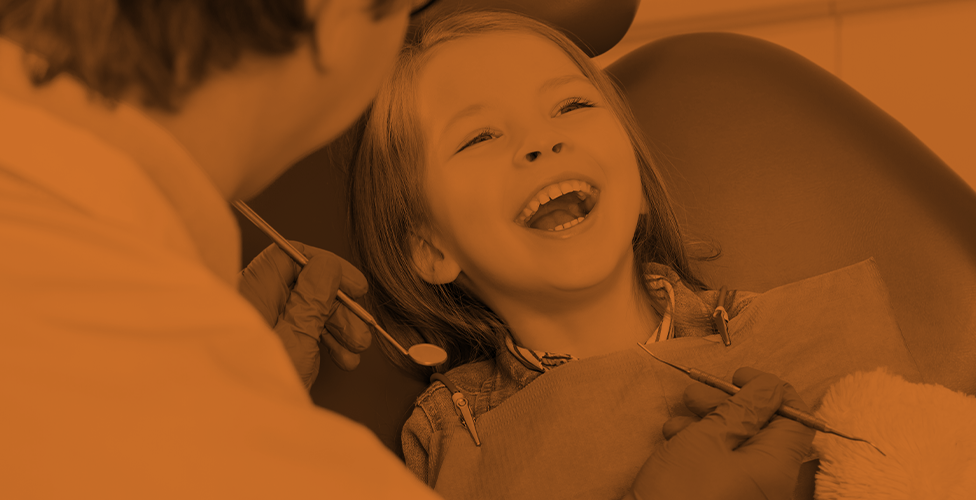 orange-scale image of a child in a dentist chair with a big smile and the back of a dentist in the foreground